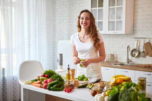 Young happy woman preparing tasty salad in the beautiful kitchen with green fresh ingredients indoors. Healthy food and Dieting concept. Loosing Weight