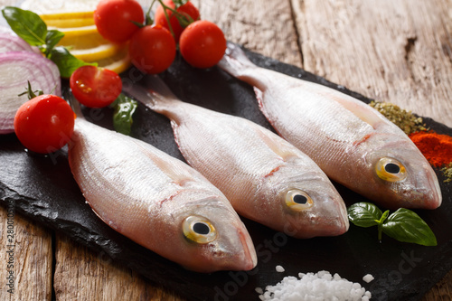 Uncooked dorado or gilt-head sea bream fish with spices, tomato, onion and lemon close up on a slate board. horizontal photo