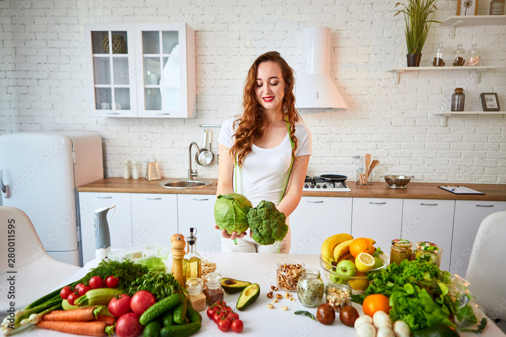 Young happy woman holding broccoli and cabbage in the beautiful kitchen with green fresh ingredients indoors. Healthy food and Dieting concept. Loosing Weight