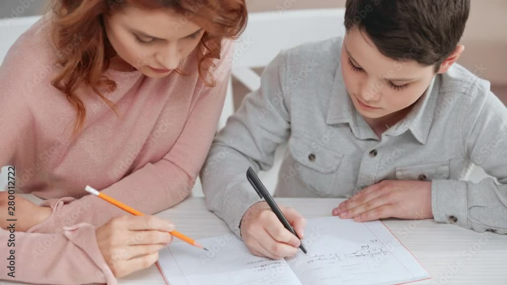 mother holding pencil and helping son with homework while he writing on copy book at desk mother holding pencil and helping son with homework while he writing on copy book at desk