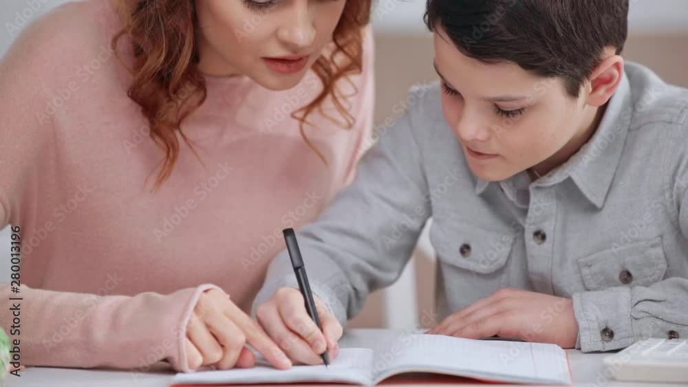 cropped view of mother helping son with homework while he writing on copy book at desk