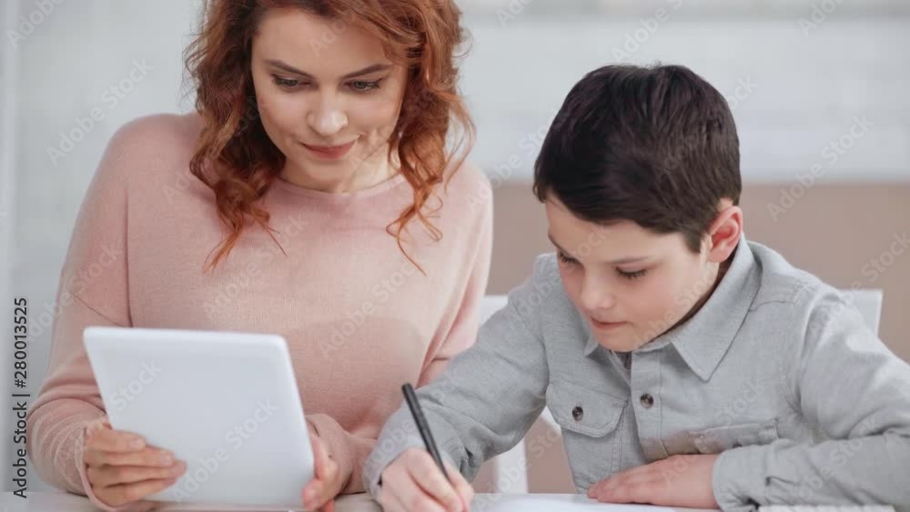 smiling mom showing digital tablet screen to son while he writing in copy book smiling mom showing digital tablet screen to son while he writing in copy book