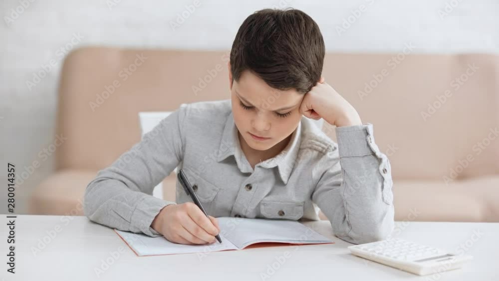 focused preteen schoolboy propping face with hand while writing in copy book at desk focused preteen schoolboy propping face with hand while writing in copy book at desk
