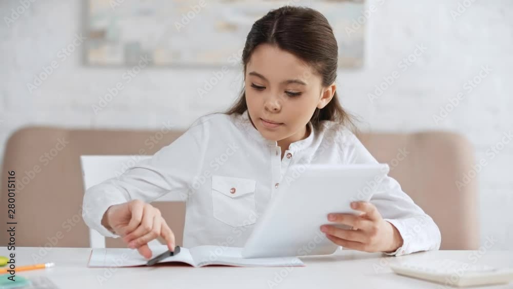 focused preteen schoolgirl using digital tablet and writing in copy book while doing homework at desk focused preteen schoolgirl using digital tablet and writing in copy book while doing homework at desk