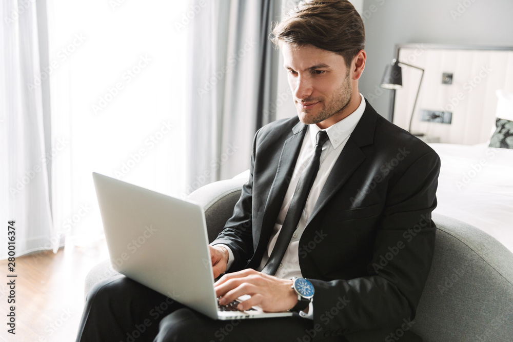 Image of confident young man sitting on sofa with laptop computer in hotel apartment during business trip Image of confident young man sitting on sofa with laptop computer in hotel apartment during business trip