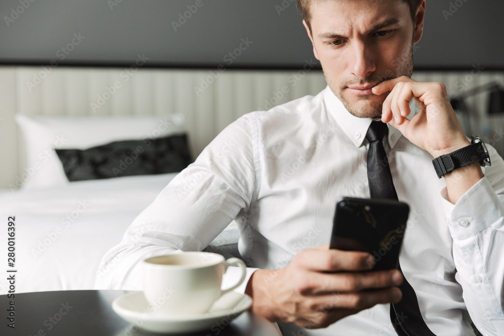 Image of serious young man holding smartphone and drinking coffee in hotel apartment during business trip Image of serious young man holding smartphone and drinking coffee in hotel apartment during business trip