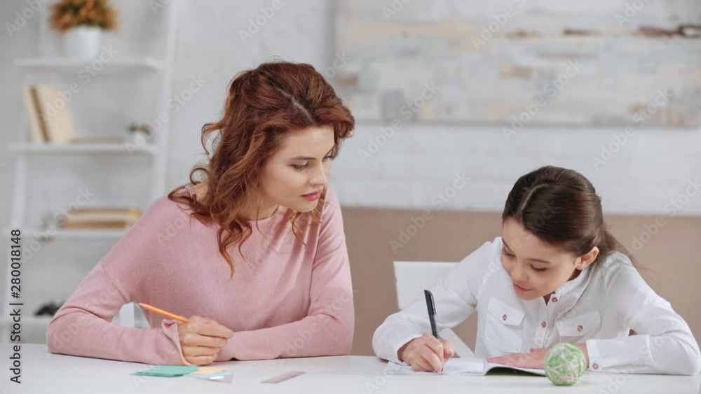 mother holding pencil and helping daughter with homework while she writing in copy book mother holding pencil and helping daughter with homework while she writing in copy book