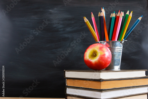 Red apple and colored pencils on stacked books. Classroom blackboard with copy space in background. Education, back to school concept