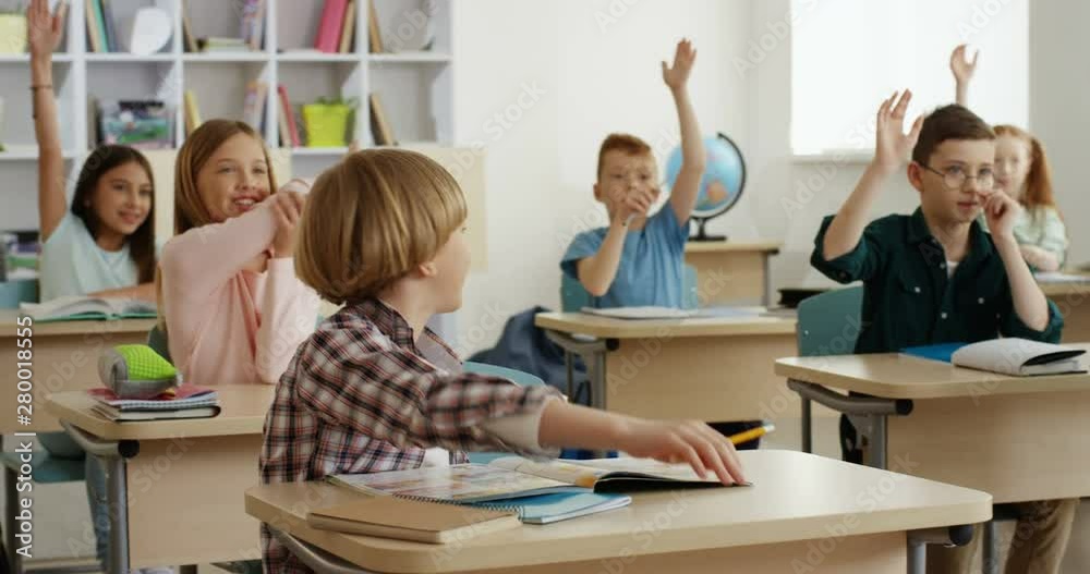 Caucasian cheerful small teen pupils at the lesson in school rising hands to answer the question of the teacher and one boy answering.