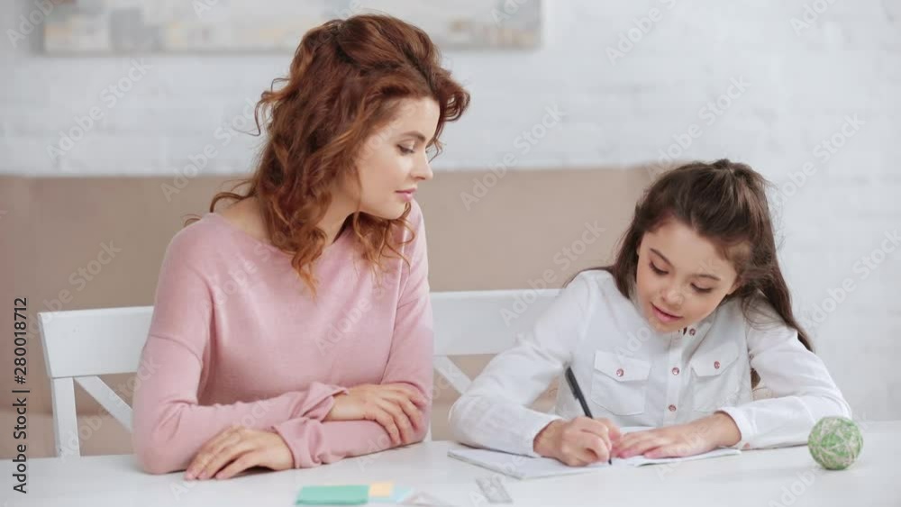 smiling mother helping daughter with homework while she writing in copy book at desk smiling mother helping daughter with homework while she writing in copy book at desk