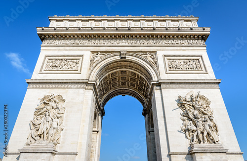 Front view of the eastern facade of the Arc de Triomphe in Paris, France, illuminated by the morning sunlight under a blue sky.