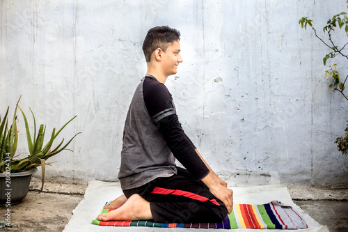 Teenage boy wearing black colored attire and doing yoga on the colorful traditional mat and doing famous Vajrasana or Diamond Pose.Horizontal isolated shot of the boy. photo