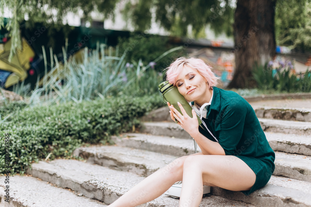 Young cheerful blondie woman listening to music, drinking water outdoors, listetning audio in earphones outdoors in green summer park. Technology, nature, healthy lifestyle, leisure concept
