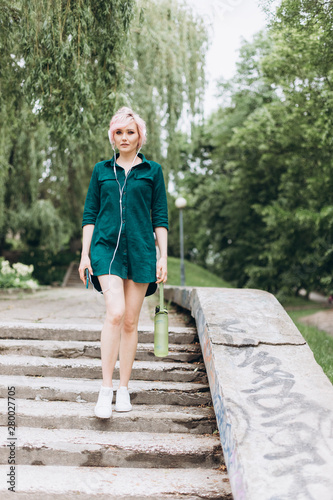 Young cheerful blondie woman listening to music, drinking water outdoors, listetning audio in earphones outdoors in green summer park. Technology, nature, healthy lifestyle, leisure concept