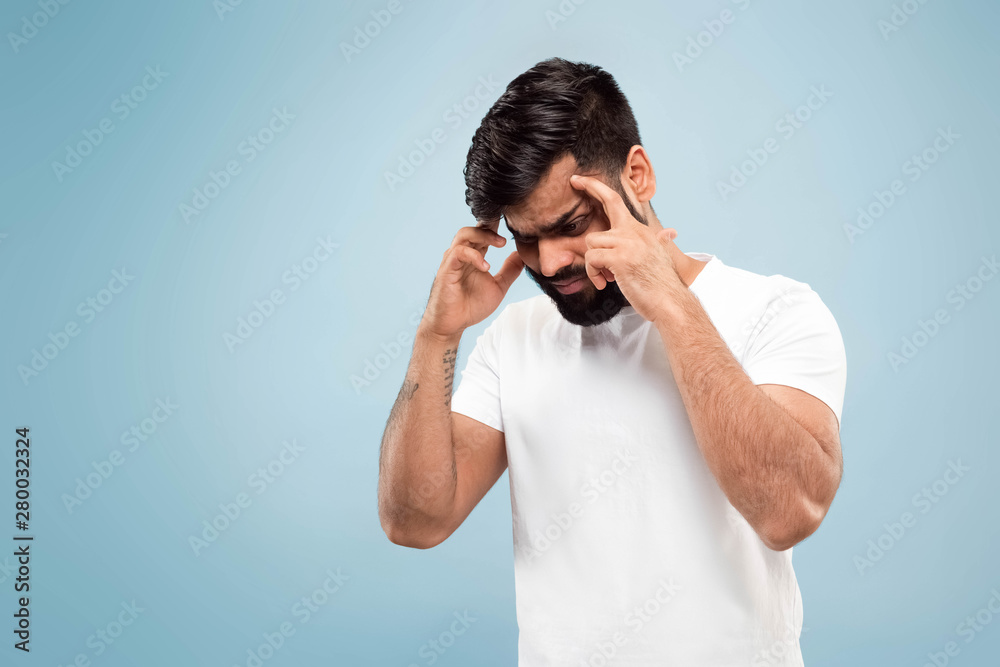 Half-length close up portrait of young hindoo man in white shirt on blue background. Human emotions, facial expression, ad concept. Negative space. Concentrating, suffering from headache. Half-length close up portrait of young hindoo man in white shirt on blue background. Human emotions, facial expression, ad concept. Negative space. Concentrating, suffering from headache.