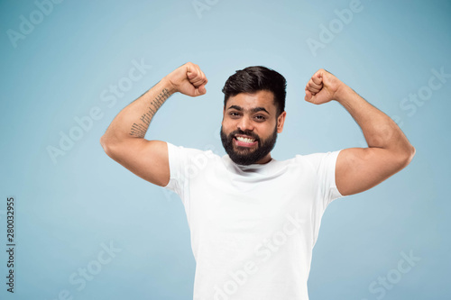 Half-length close up portrait of young hindoo man in white shirt on blue background. Human emotions, facial expression, ad concept. Negative space. Celebrating, winning, crazy happy.