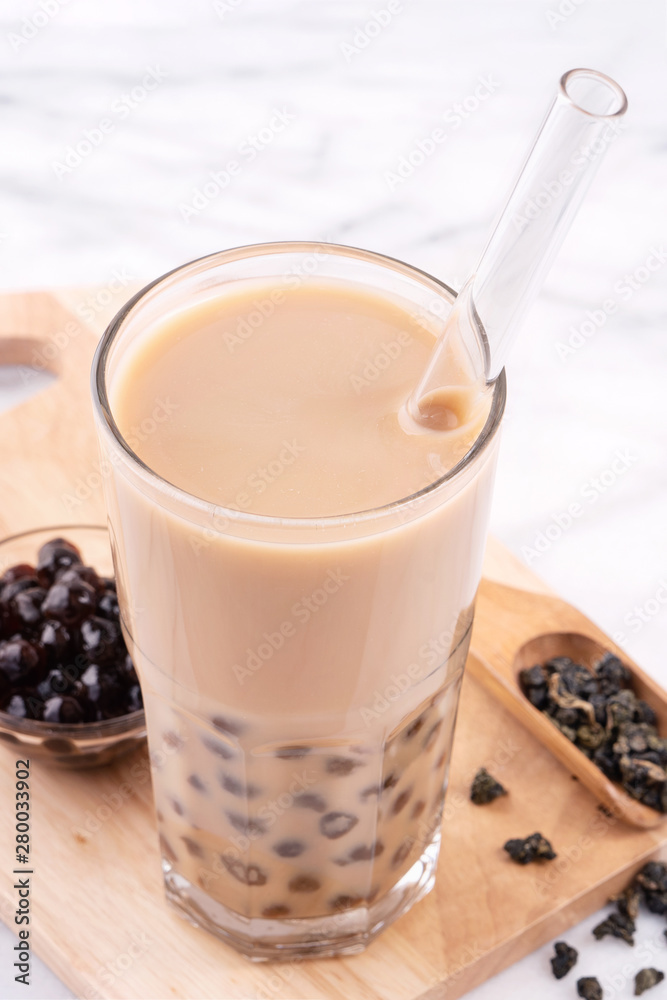 Tapioca pearl ball bubble milk tea, popular Taiwan drink, in drinking glass with straw on marble white table and wooden tray, close up, copy space. Tapioca pearl ball bubble milk tea, popular Taiwan drink, in drinking glass with straw on marble white table and wooden tray, close up, copy space.