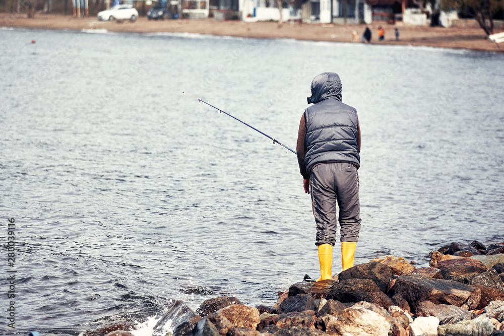 Male fisherman standing on the rocks near the seaside and fishing on a cloudy winter day Male fisherman standing on the rocks near the seaside and fishing on a cloudy winter day