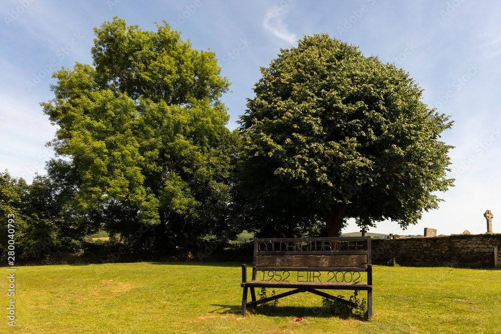 An old bench on a street in the center of the town of Whitecombe in the English county of Devon in Dartmoor. In the background are beautiful trees in the sunshine. An old bench on a street in the center of the town of Whitecombe in the English county of Devon in Dartmoor. In the background are beautiful trees in the sunshine.