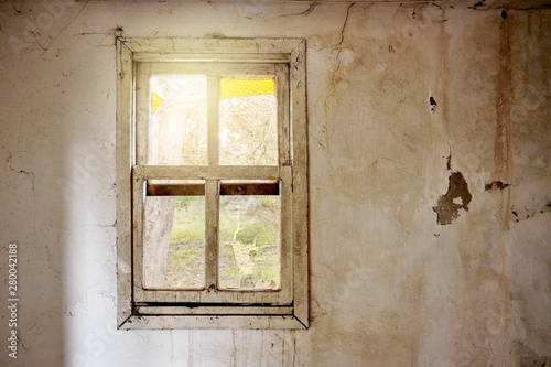 Interior of a ruined house with old, dirty and cracked white wall and a broken window frame with a green meadow field view