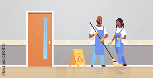 couple cleaners in uniform working together cleaning service concept african american man woman janitors using mop spray plastic bottle modern clinic corridor interior full length flat