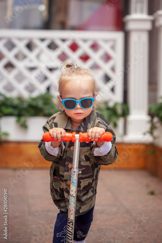 Cool stylish baby girl in sunglasses on scooter in the street. photo