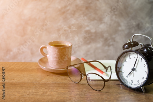 Close-up of retro alarm black clock vintage style with Blank open book, red pencil and glassescup coffee pastel pink old on wooden table. Time for Wake up early. light morning through the window.