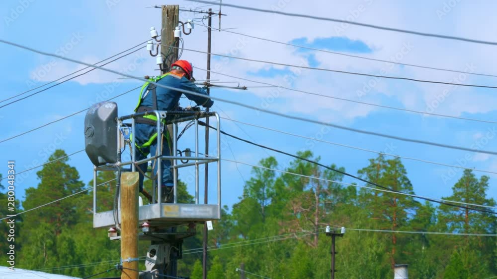 Electrician in uniform repairs power lines, standing on the bucket. Summer day. In the background is a blue cloudy sky.
