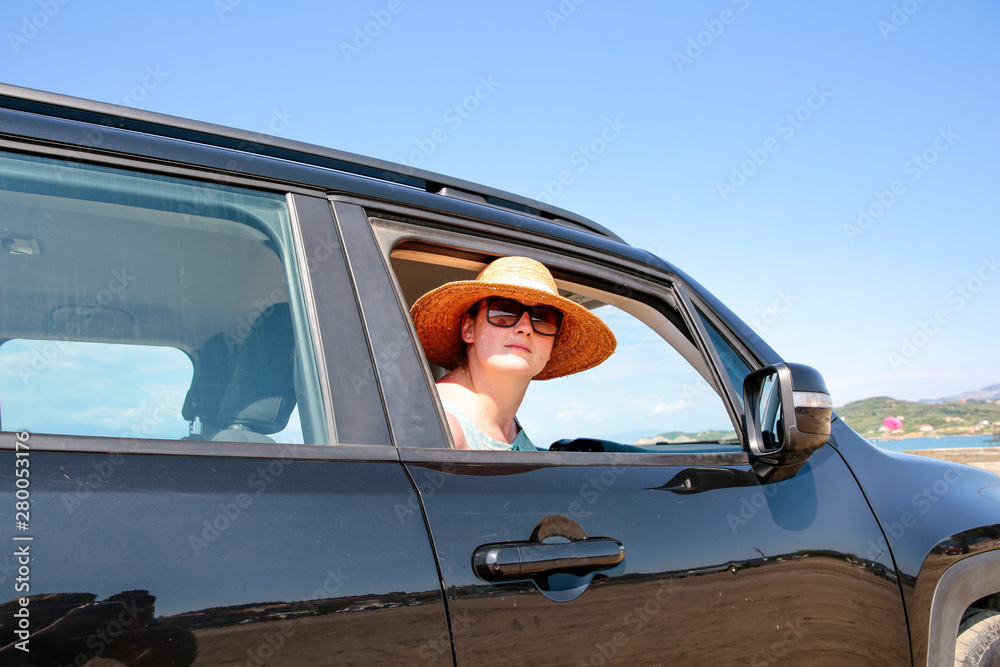 Naklejka premium A woman weraing sunglasses and a hat in the summer car on the beach and ocean view..
