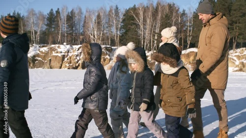 Tracking medium shot of happy devoted father and mother hiking with their five young kids outdoors in snow over frozen quarry with stony slopes, in soft afternoon glow, admiring views and chatting
