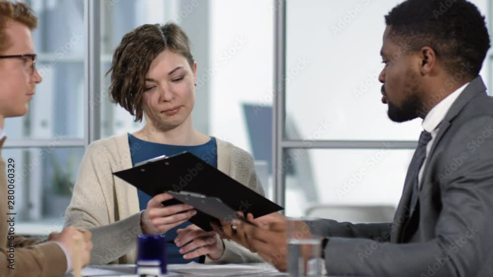 Tracking waist-up shot of African American male solicitor, wearing business suit and tie, consulting young Caucasian couple in his office and giving them draft copies of prenuptial agreement Tracking waist-up shot of African American male solicitor, wearing business suit and tie, consulting young Caucasian couple in his office and giving them draft copies of prenuptial agreement