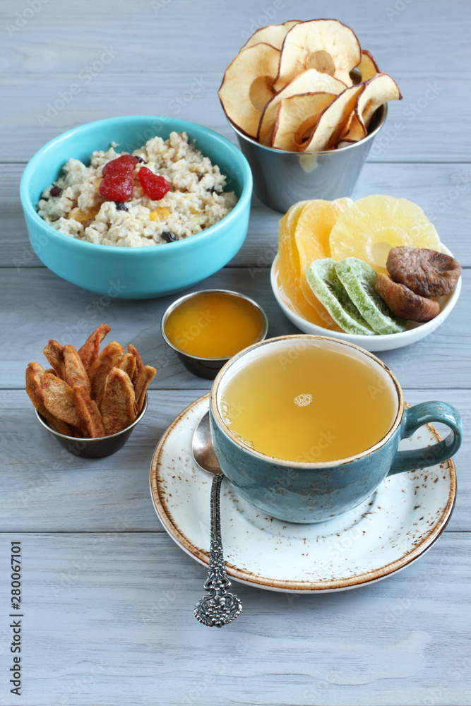 Oatmeal with fruit pieces and candied strawberries, pieces of candied pineapple, kiwi and mango, dried figs, apple and pear chips, honey in small bowls and a cup of green tea on a gray wooden table. Oatmeal with fruit pieces and candied strawberries, pieces of candied pineapple, kiwi and mango, dried figs, apple and pear chips, honey in small bowls and a cup of green tea on a gray wooden table.