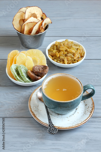 Vegetarian dessert: pieces of candied pineapple, mango and ginger root, dried figs, golden raisins, apple chips in small bowls and a cup of green tea on a gray wooden table. Closeup