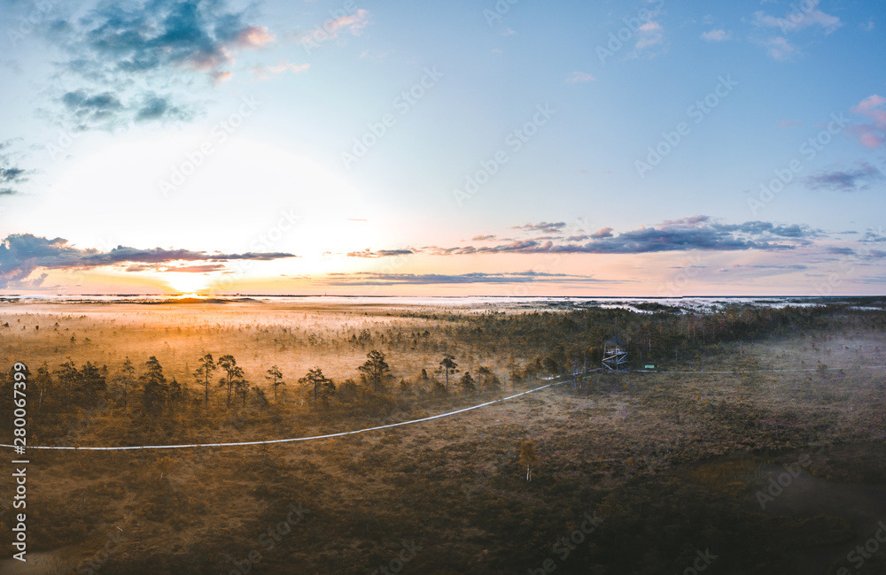  Warmly colored sunrise over a foggy swamp. Aerial view of stunning landscape at peat bog at Cenas Tirelis in Latvia. Wooden trail leading along the lake surrounded by pounds and forest. 