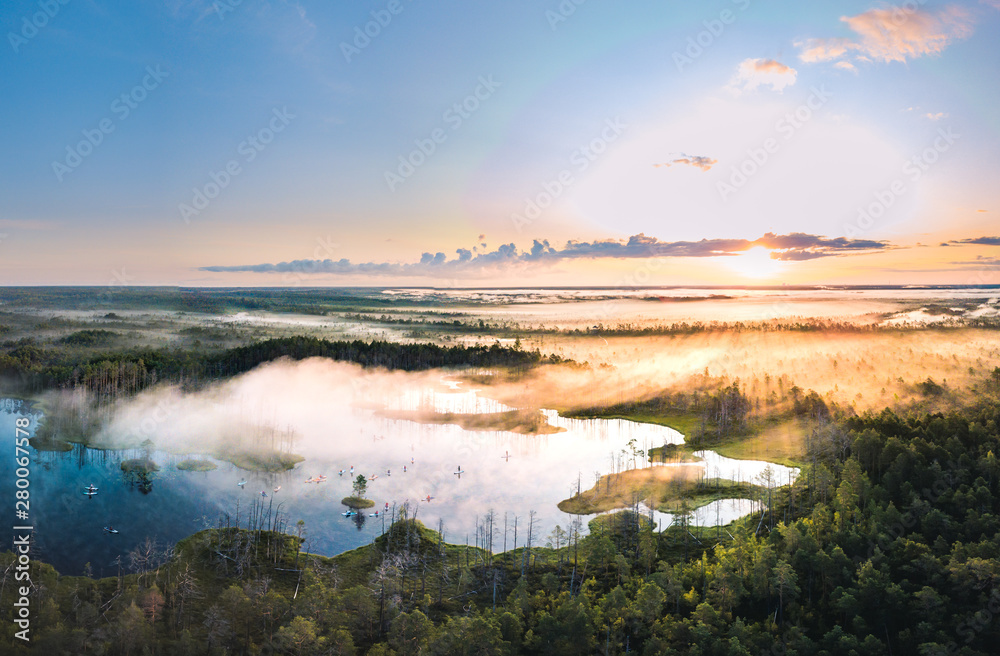  Warmly colored sunrise over a foggy swamp. Aerial view of stunning landscape at peat bog at Cenas Tirelis in Latvia. Wooden trail leading along the lake surrounded by pounds and forest. 