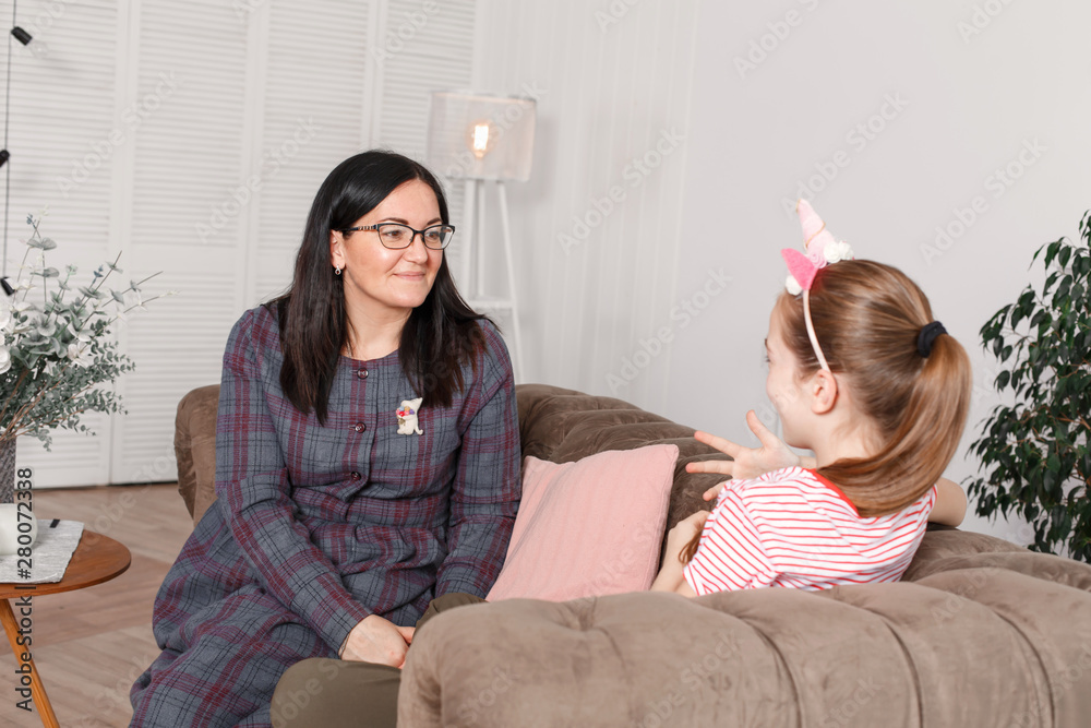 Mom and daughter are sitting on the couch and chatting. Girl teenager with emotions tells her mother a story. Daughter shares her feelings with her parent. Leisure mothers and daughters