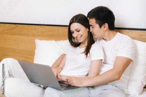 Happy young couple of guy and girl is lying in bed and watching film on laptop. Young married couple lying on bed at home and using laptop. Happy couple is lounging in bed before going to bed