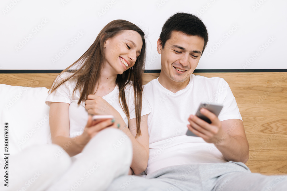 Attractive young married couple is sitting on sofa in living room and laughs watching video on smartphone. Portrait of happy loving beautiful couple sitting on bed in bright modern apartment Attractive young married couple is sitting on sofa in living room and laughs watching video on smartphone. Portrait of happy loving beautiful couple sitting on bed in bright modern apartment