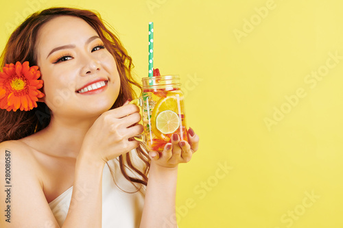 Portrait of Asian beautiful girl with flower in her red hair smiling at camera while drinking lemonade with slices of orange and lemon