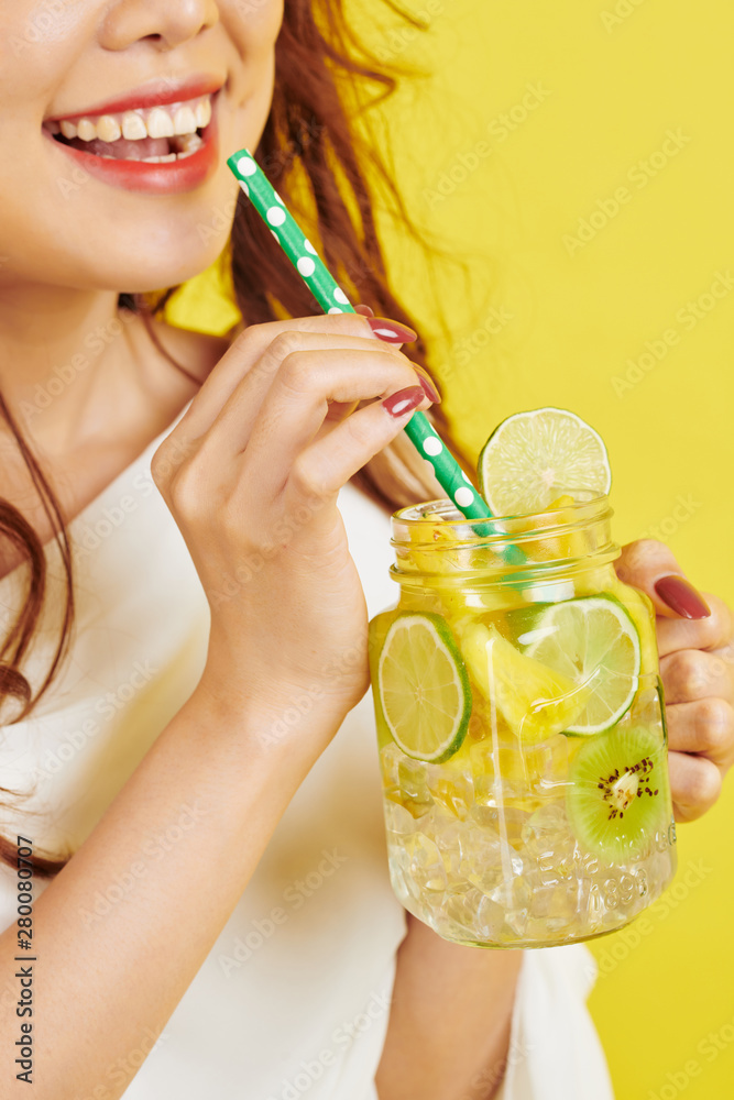 Fototapeta premium Close-up of happy young girl holding glass with cold lemonade with slices of lemon and drinking it from drinking straw isolated on yellow background