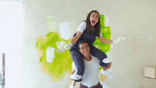Couple is having fun redecorating their new appartment. Happy girl sits on the shoulders of the guy and paints the wall paint