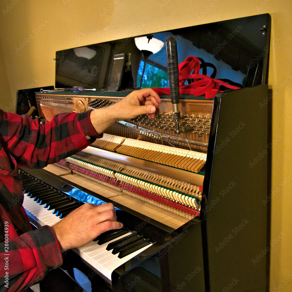 Technician tuning an upright piano by playing notes on black and white keys and using lever and tools to tighten or loosen the strings to produce proper pitch.