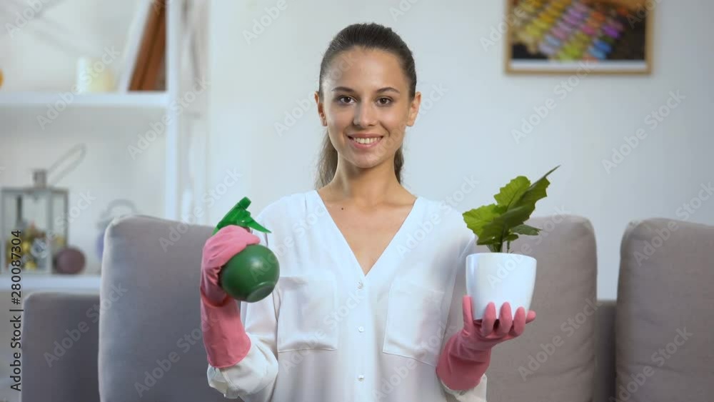 Smiling woman holding water sprayer and flowerpot, looking at camera, home Smiling woman holding water sprayer and flowerpot, looking at camera, home