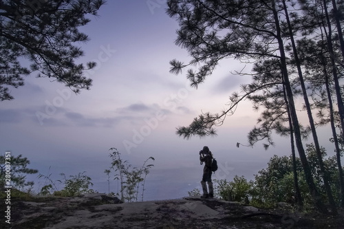 Silhouette a Photographer shooting photo under pine trees with cloudy sky background, sunrise at Nok Aen Cliff, Phu Kradueng, Loei, Thailand. © Yuttana Joe