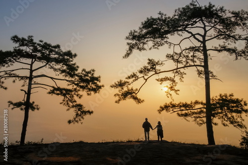 Mountain view Silhouette couple tourists standing on top hill under pine tree with yellow sun light in the sky background  sunrise at Nok Aen Cliff  Phu Kradueng  Loei  Thailand.