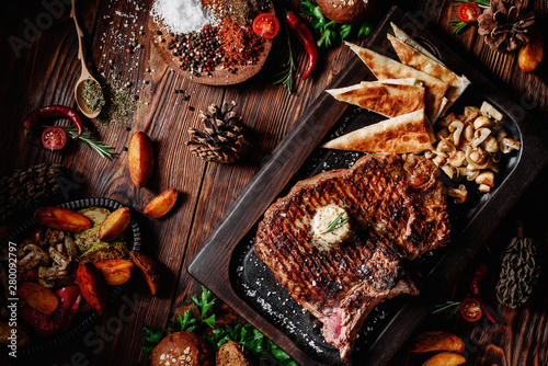 Juicy grilled steak T-Bone decorated rosemary on dark board with mushrooms and fried pita bread on dark wooden background in beautiful composition among vegetables and spices. Top view. Flat lay