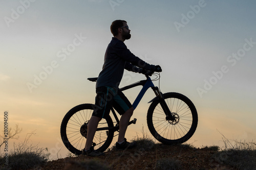Cyclist in shorts and jersey on a modern carbon hardtail bike with an air suspension fork rides off-road on the orange-red hills at sunset evening in summer 