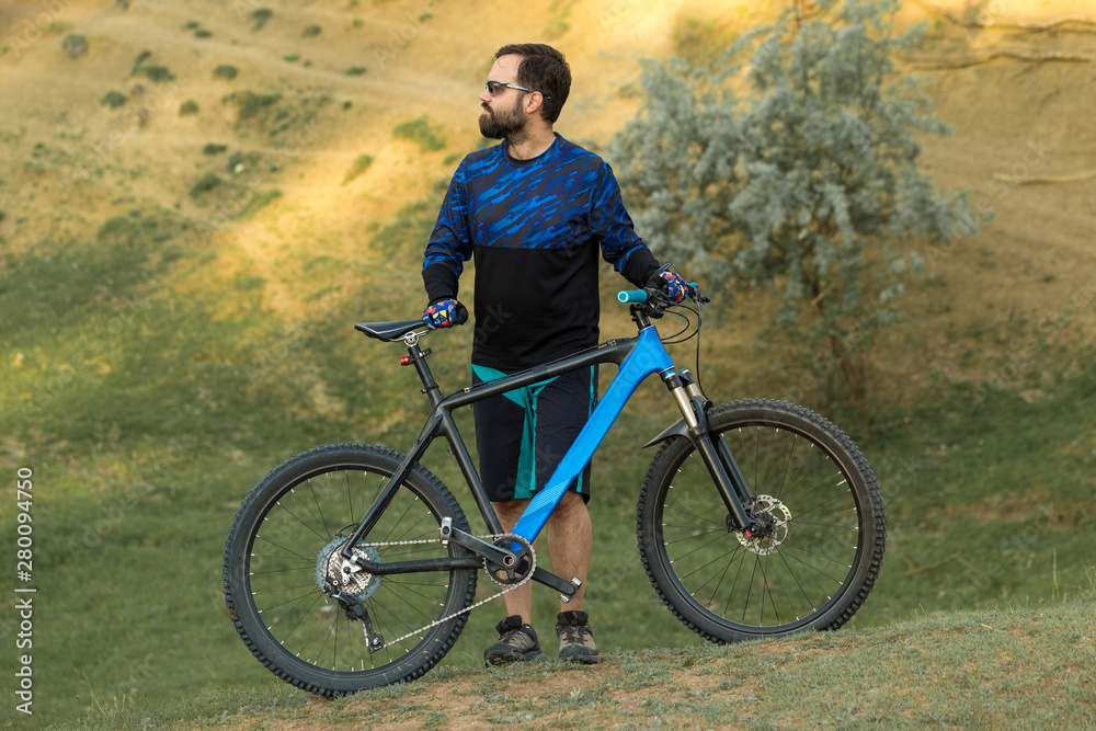 Cyclist in shorts and jersey on a modern carbon hardtail bike with an air suspension fork rides off-road on the orange-red hills at sunset evening in summer Cyclist in shorts and jersey on a modern carbon hardtail bike with an air suspension fork rides off-road on the orange-red hills at sunset evening in summer