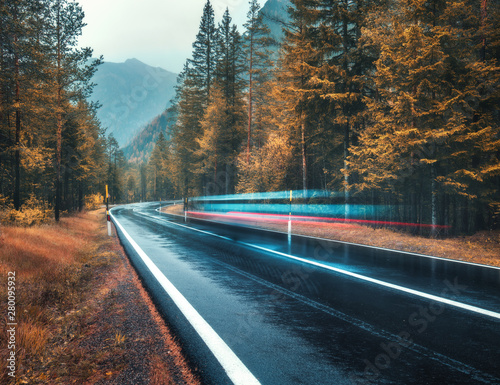 Blurred car on the road in autumn forest in rain. Perfect asphalt mountain road in overcast rainy day. Roadway, orange trees in alps in fall. Nature. Highway in foggy woodland. Car in motion. Travel photo