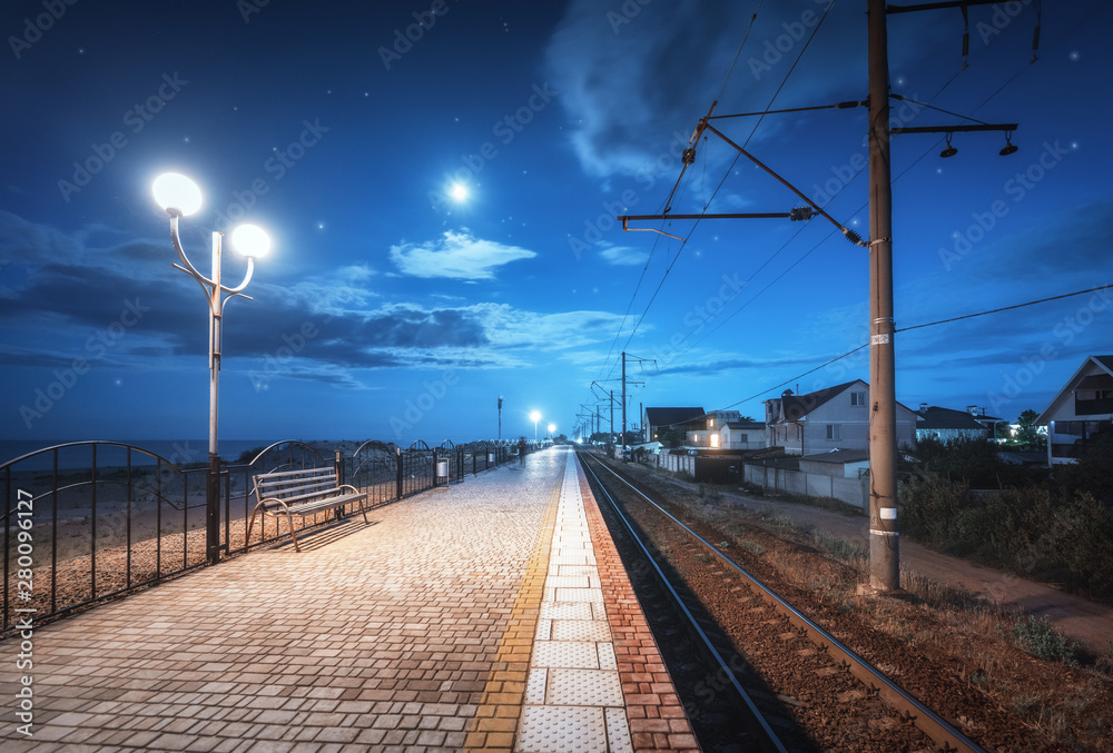 Beautiful railway station at night in summer. Starry sky over railroad at dusk. Industrial landscape with railroad, railway platform, city lights, buildings, blue sky with moon and clouds at twilight